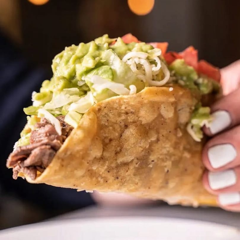 Hand holding a crispy El Taquito taco filled with meat, guacamole, lettuce, and tomatoes.