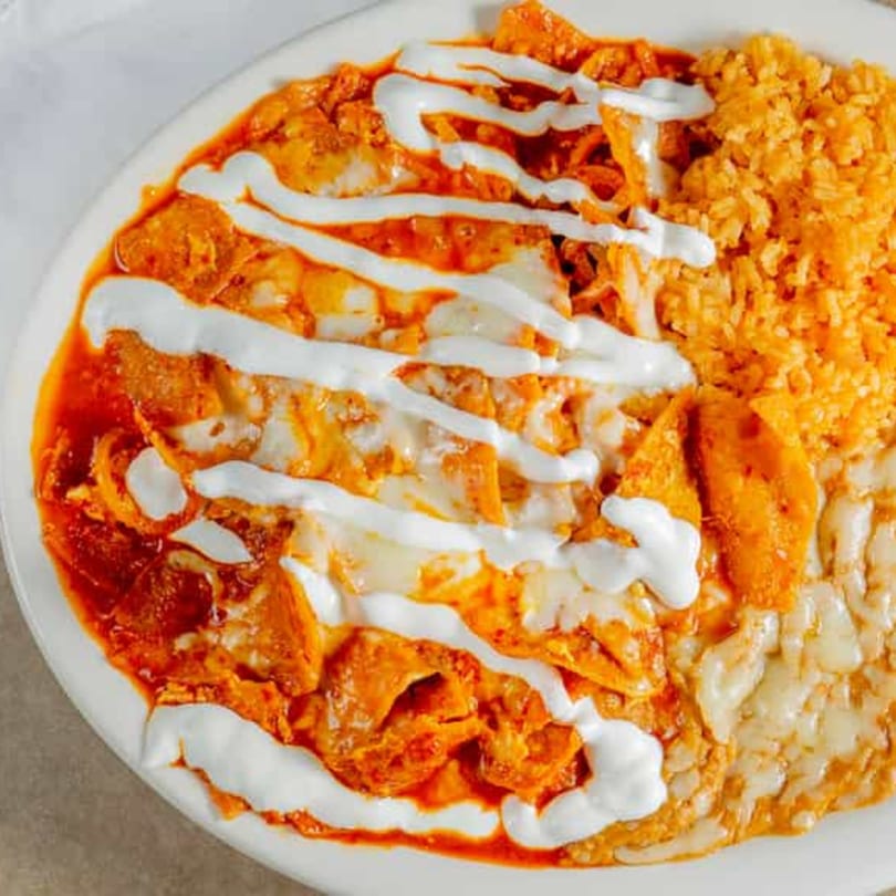 Chilaquiles with red sauce, melted cheese, sour cream, rice, and refried beans on a white plate.