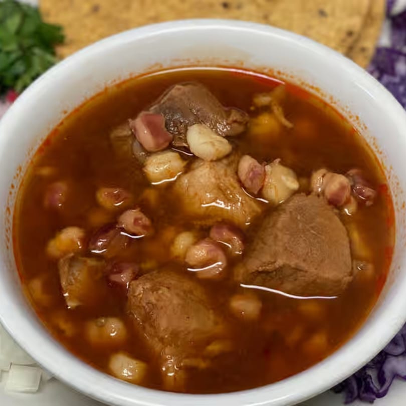 A white bowl filled with El Taquito Restaurant Dos's pozole, a rich red Mexican soup with meat and hominy.