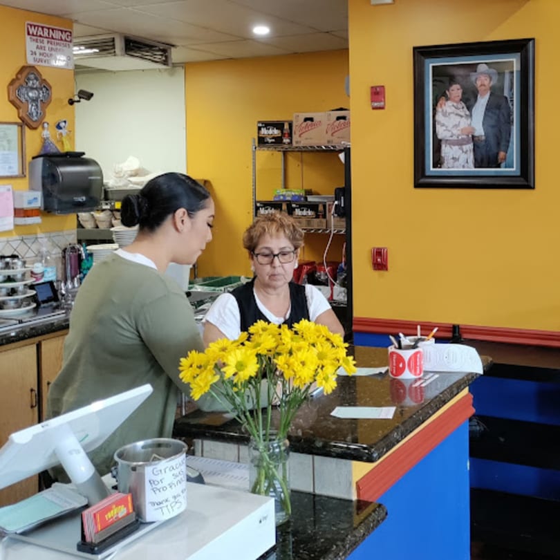 Two women at the counter of El Taquito Restaurant Dos, with yellow walls and a framed photo.
