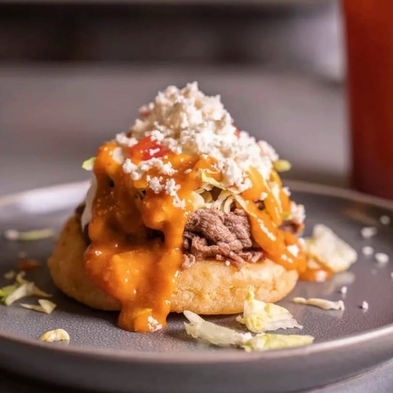 A Mexican sopes dish with shredded beef, orange sauce, crumbled white cheese, and lettuce.