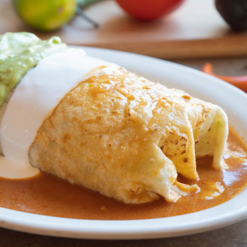A large, saucy burrito topped with sour cream and guacamole on a white plate.