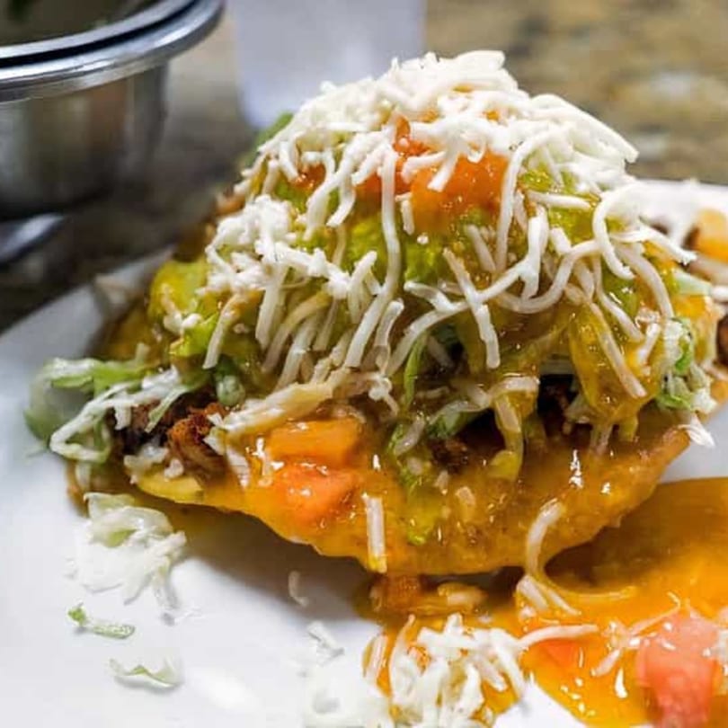 A close-up of a Mexican tostada topped with shredded cheese, lettuce, and salsa on a white plate.